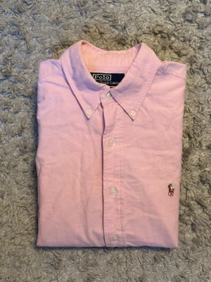 Rosa skjorta från Polo Ralph Lauren - Klassisk rosa skjorta från Polo Ralph Lauren med button-down krage och broderad logga på bröstet. Skjortan har långa ärmar, vita knappar och är tillverkad i mjuk bomull. Perfekt för en stilren och avslappnad look.