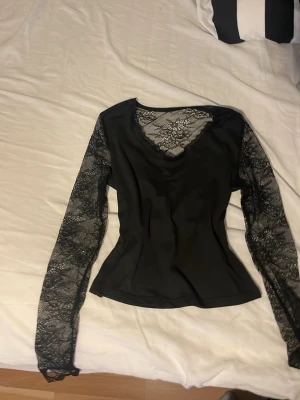 Svart topp med spetsdetaljer - Snygg svart topp med långa spetsärmar och spets vid ringningen. Figurnära passform och transparenta detaljer på ärmarna ger en elegant look. Perfekt för dig som vill ha något extra till din outfit.
