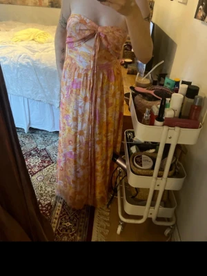 Blommig långklänning med rosett - Säljer en somrig långklänning med blommigt mönster i orange, rosa och vitt. Klänningen är axelbandslös med en stor rosett framtill och har en flowy passform. Perfekt för varma dagar och midsommarfirande.