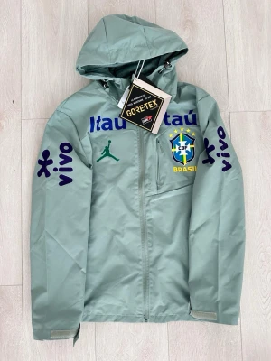 Brasil vindjacka Jordan x CBF - Ljusgrön vindjacka med huva från Jordan i samarbete med Brasiliens fotbollslandslag (CBF). Jackan har GORE-TEX-material, tryckta loggor i blått, grönt och gult samt bröstficka med dragkedja. Snygga detaljer med 'vivo' på ärmarna och justerbara muddar.