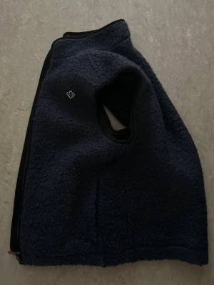 Mörkblå fleeceväst med dragkedja - En mörkblå skön väst i mjuk fleece med dragkedja längs sidan och öppning för armarna. Västen har en enkel design med en liten broderad detalj på bröstet. Perfekt för lager-på-lager och håller dig varm under kyliga dagar men även under vår/sommar kvällar! 
