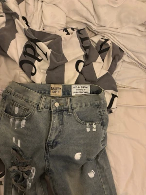 Distressed jeans från Gallery Dept. - Säljer ett par gråblå jeans från Gallery Dept. med kraftigt slitna detaljer och vita färgstänk. Jeansen har klassisk femficksmodell och raka ben. Perfekta för dig som gillar en edgy och unik streetwear-look.