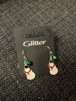 Snögubbeörhängen från Glitter - Ett par söta örhängen från Glitter med snögubbar i vitt och svart, dekorerade med röda halsdukar och gröna pärlor. Perfekta för dig som vill ha en lekfull och julig accessoar. Krokar i metall.
