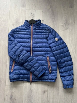 Daniel - Blå dunjacka från Moncler. En liten laglapp på ryggen som inte påverkar helhetsintryck eller syns vid användning. Storlek 3 sitter som en M.