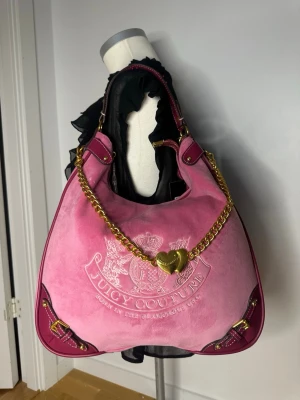 Juicy Couture rosa axelväska i sammet - daydreamer - Stor rosa handväska från Juicy Couture i mjuk sammet med broderad logga framtill. Väskan har detaljer i mörkrosa läder, guldfärgad kedja med hjärtberlock och dekorativa spännen i hörnen. Perfekt för dig som gillar statement-accessoarer. Mått: Bredd: 34 cm, Längd: 28 cm och Djup: 8 cm