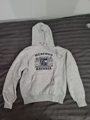 Grå München Bavaria hoodie - Snygg grå hoodie från Fruit of the Loom med tryck 'München Bavaria' och ett lejonmotiv på bröstet. Klassisk modell med huva och dragsko samt ribbade muddar vid ärmslut och midja. Perfekt för en chill och avslappnad stil.