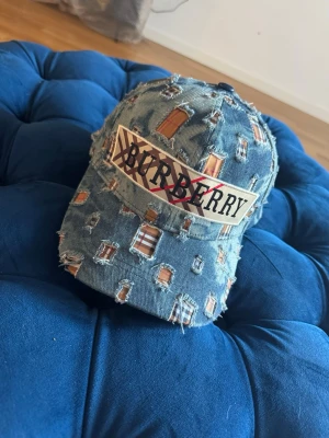 Burberry denim keps med slitningar - Säljer en blå denim keps från Burberry med coola slitningar och ett stort Burberry-tryck framtill. Kepsen har en klassisk böjd skärm och justerbar passform. Perfekt för dig som gillar streetstyle och vill sticka ut med en unik accessoar.