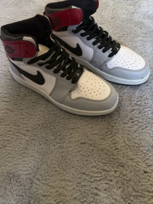 Nike Air Jordan 1 High vit/röd/svart - Säljer ett par Nike Air Jordan 1 High sneakers i vitt, grått, svart och rött. Klassisk hög modell med svart swoosh, perforerad tåbox och svarta skosnören. Tillverkade i läder med ikoniska detaljer på ankeln och tungan.
