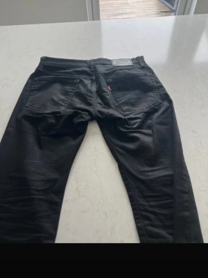 Svarta Levi's jeans raka ben - Säljer ett par svarta jeans 502 från Levi's med klassisk femficksdesign och raka ben. Jeansen har en diskret röd Levi's-tag på bakfickan och är tillverkade i ett slitstarkt bomullsmaterial. Perfekta för en clean och tidlös look.