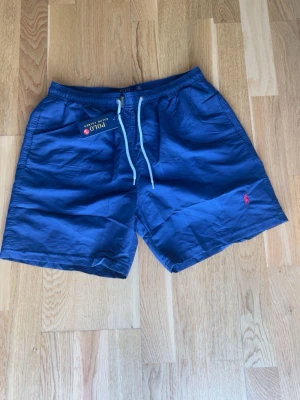 Blå shorts från Polo Ralph Lauren - Snygga marinblå shorts från Polo Ralph Lauren med dragsko i midjan och klassisk röd logga broderad på ena benet. Shortsen har en avslappnad passform och är perfekta för sommaren. Tillverkade i ett lätt och bekvämt material.