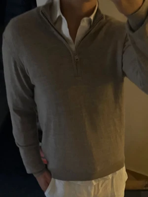 Beige halvzip-tröja från Oscar Jacobson  - Snygg beige långärmad tröja från oscar Jacobson med halvzip och ribbad krage. Perfekt att bära över en skjorta för en clean och stilren look. Tillverkad i mjukt material som känns skönt mot huden. 100% merinoull