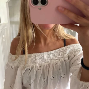Vit offshoulder blus med broderi - Snygg vit offshoulder blus med broderade hålmönster och smock upptill. Blusen har vida långa ärmar och en romantisk känsla med vågig kant nertill. Perfekt till jeans för en trendig look.
