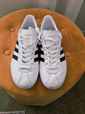 Adidas Originals vita sneakers st 38 - Klassiska Adidas Originals sneakers i vitt läder med svarta ränder på sidorna och guldfärgade detaljer på plösen och hälen. Låg modell med vit sula och svart hälparti. Perfekta för en clean och sportig stil.