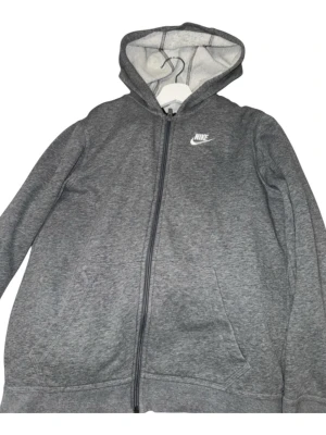 Grå zip-hoodie från Nike - Grå hoodie från Nike med dragkedja och vit broderad logga på bröstet. Mjuk insida, huva med dragsko och två fickor framtill. Perfekt för en avslappnad och sportig stil.