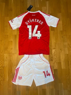 Arsenal matchställ Gyökeres #14 - Säljer ett Arsenal matchställ med Gyökeres och nummer 14. Tröjan är röd med vita ärmar och detaljer, Adidas-logga och Emirates Fly Better-tryck. Shortsen är vita med röda ränder och Arsenal-emblem. Perfekt för fotbollsträning eller match.