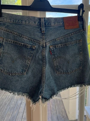 Levi's jeansshorts med fransig kant - Klassiska blå jeansshorts från Levi's med rå, fransig kant och femficksmodell. Shortsen har hög midja och är tillverkade i slitstark bomull. Perfekta för sommardagar.