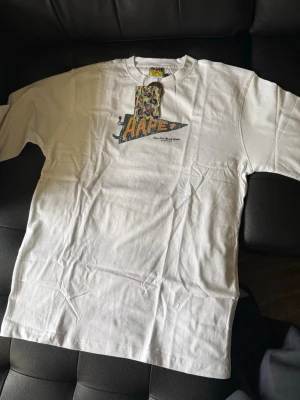 Vit AAPE t-shirt med camo-detalj - Snygg vit t-shirt från AAPE by A Bathing Ape med tryck på bröstet och camo-detalj i grönt, lila och beige. Klassisk passform och rund halsringning. Perfekt för dig som gillar streetwear och vill sticka ut med en ikonisk logga.