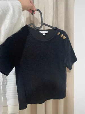 Svart topp med guldknappar - Stilren svart topp med korta ärmar och rund hals. Toppen har tre dekorativa guldknappar på ena axeln som ger en snygg detalj. Materialet ser ut att vara mjukt och lite fluffigt, vilket gör den extra mysig och trendig.