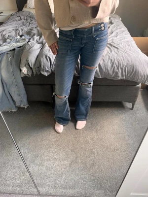 Gina 14+ jeans - Säljer ett par blå bootcut jeans med slitningar och stora hål vid knäna. Jeansen har låg midja, dubbla knappar i linningen och fem fickor. Perfekta för en avslappnad och trendig look.
