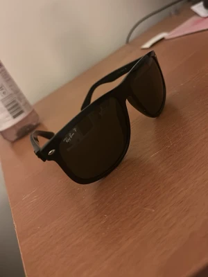 Svarta Ray-Ban Wayfarer solglasögon - Snygga svarta/mörkgrön Ray-Ban Wayfarer solglasögon med klassisk fyrkantig form och mörka glas. Perfekta för att ge din stil en cool touch. Ikonisk logga på linsen och robust plastbåge.