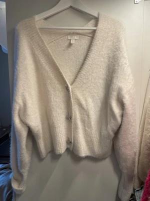 Vit fluffig kofta med pärlknappar - Mysig vit kofta med fluffig textur och djup V-ringning. Tre dekorativa pärlknappar framtill och ribbade muddar vid ärmslut och nederkant. Perfekt för lager på lager och ger en soft look till din outfit.