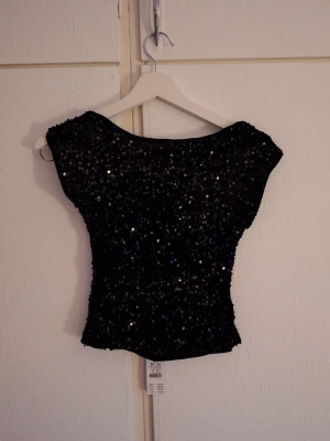 Svart glittrig topp med paljetter - Topp i paljett med öppen rygg. Toppen är svart och har båtringning. Den har en ärmlös design, en kortare längd och en figurnära passform. Helt ny med prislapp.