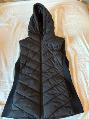 Svart dunväst med huva - Snygg svart dunväst med dragkedja framtill och stor, vadderad huva. Västen har ett quiltat mönster i diagonala ränder och är något längre i modellen. Perfekt för lager-på-lager och streetwear-stil. Passar både S och M