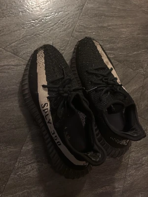 Yeezy Boost 350 V2 Black/White - Säljer ett par Adidas Yeezy Boost 350 V2 i svart med vit stripe och texten SPLY-350 på sidan. Skorna har en stickad ovandel i Primeknit och en chunky, räfflad sula i gummi. Ikonisk siluett som är superpopulär bland sneakerheads.
