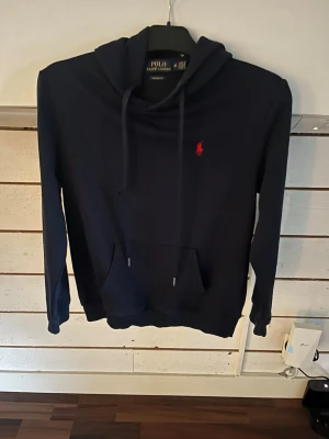 Mörkblå hoodie från Polo Ralph Lauren - Snygg mörkblå hoodie från Polo Ralph Lauren med klassisk röd logga på bröstet. Hoodien har huva med dragsko och en stor magficka framtill. Tillverkad i mjukt material och har ribbade muddar vid ärmar och nederkant. Perfekt för en chill och stilren look, den är också custom fit
