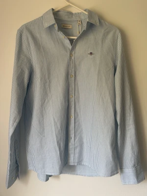 Blårandig skjorta från Gant - Klassisk blårandig skjorta från Gant med lång ärm och knappar framtill. Skjortan har en broderad logga på bröstet och är tillverkad i bomull. Perfekt för en stilren och avslappnad look.