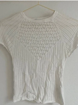 Vit stickad topp med spetsdetaljer - Snygg vit stickad topp med korta ärmar och spetsmönster framtill. Perfekt för dig som gillar en luftig och feminin stil. Toppen har rund halsringning och är lätt att matcha med jeans eller kjol.