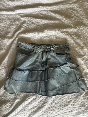 Volangkjol i denim med jeanslook - Snygg kort jeanskjol i ljusblå denim med volanger och råa kanter. Klassisk femficksmodell med bälteshällor och knappstängning framtill. Perfekt för dig som gillar en trendig och avslappnad stil.