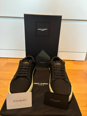 Saint Laurent sneakers - Svarta sneakers från Saint Laurent i canvas med vit broderad logga på sidan och gulddetaljer på plösen. Skorna har vita gummisulor och klassisk låg profil. Perfekta för en clean och stilren look.