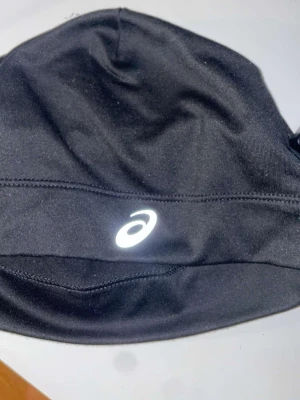 Svart sportmössa från Asics - Säljer en svart sportmössa från Asics med reflekterande logga framtill. Mössan är tillverkad i ett stretchigt och funktionellt material som passar perfekt för träning utomhus. Snygg och enkel design som håller dig varm under kalla dagar.