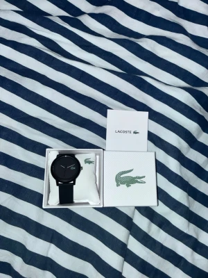 Svart Lacoste klocka i originalask - Snygg svart klocka från Lacoste med minimalistisk design och diskret Lacoste-logga på urtavlan. Klockan har svart armband och levereras i originalask med kudde och logotyp. Perfekt accessoar för en stilren look. Pris går att diskutera.