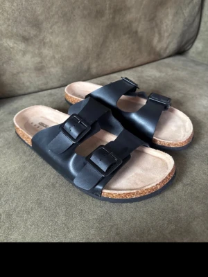 Svarta sandaler med dubbla spännen - Stilrena svarta sandaler med två breda remmar och justerbara spännen. Fotbädden är ljusbeige och sulan har en klassisk korklook. Perfekta för en avslappnad och trendig stil. Passar till både jeans och shorts.
