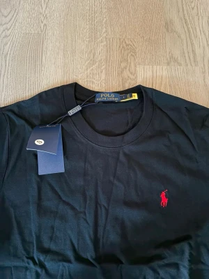 Svart t-shirt från Polo Ralph Lauren - Klassisk svart t-shirt från Polo Ralph Lauren med röd broderad logga på bröstet. T-shirten har rund halsringning och korta ärmar. Tillverkad i mjuk bomull för en skön känsla och enkel stil.