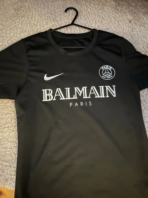 Svart Balmain x Nike Paris T-shirt - Svart t-shirt från Balmain i samarbete med Nike och Paris, med vit logga och tryck framtill. Klassisk rund hals och korta ärmar. Perfekt för dig som gillar streetwear och sportig stil.