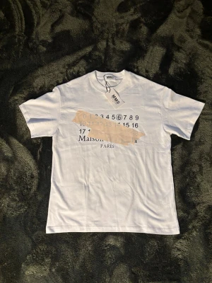 Vit Maison Margiela t-shirt - Vit t-shirt från Maison Margiela med svart tryck framtill som visar siffror och logga. Klassisk passform med rund hals och korta ärmar. Tillverkad i mjuk bomull och har en stilren look med märkets signaturdetaljer. Kom privat om du har frågor! 