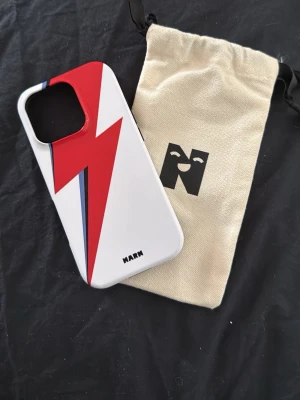 IPhone 16 pro skal från NARN - Snyggt mobilskal i hårdplast från NARM med vit bas och en röd blixt med blå detalj på baksidan. Skalet har svart insida och passar perfekt för dig som vill sticka ut med din mobil. Levereras med en beige tygpåse med logga. 