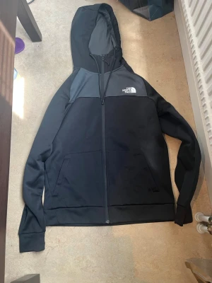 Svart hoodie från The North Face - Snygg svart hoodie från The North Face med dragkedja och två stora fickor framtill. Hoodien har en klassisk logga på bröstet och är tillverkad i ett mjukt och bekvämt material. Perfekt för en avslappnad streetstyle-look.