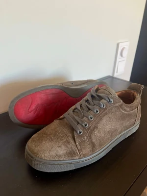 Christian Louboutin skor  - Snygga sneakers i brun mocka med grå snörning och grå sula. Skorna har en låg profil och en cool röd detalj på yttersulan. Perfekta för dig som gillar en stilren och avslappnad look.