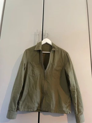 Olivgrön overshirt med dragkedja - Stilren olivgrön overshirt i bomull med två bröstfickor och dragkedja framtill. Klassisk krage och raka ärmar med knapp vid ärmslut. Perfekt lager på lager-plagg för en avslappnad look.