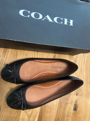 Svarta ballerinaskor från Coach - Ett par svarta ballerinaskor från Coach. Skorna har en liten rosett framtill och en platt sula. Insidan är i en ljusbrun nyans med Coach-logotypen präglad. Yttersulan är svart med ett mönster av Coach-logotypen.