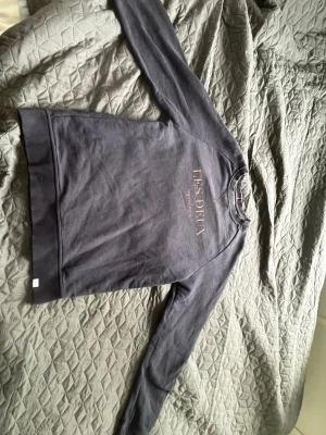 Mörkgrå sweatshirt från Les Deux - Mörkgrå sweatshirt från Les Deux med orange tryckt text på bröstet. Tröjan har rund halsringning och långa ärmar. Perfekt för en chill och stilren look.