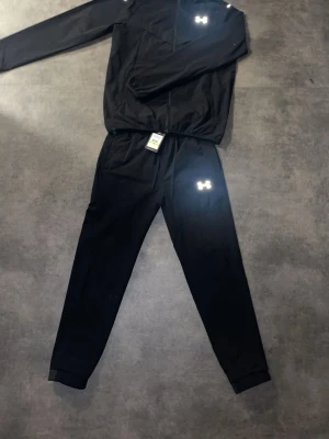Svart träningsset från Under Armour - Snyggt svart träningsset från Under Armour med jacka och byxor. Setet har reflekterande loggor på både jacka och byxa, dragkedja på jackan och resår i midjan på byxorna. Perfekt för träning eller chill. Materialet är lätt och andas.