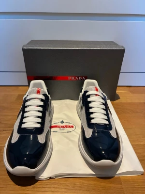 Prada skor  - Snygga sneakers från Prada i marinblått och vitt med meshpaneler och lackdetaljer. Klassisk låg modell med vita skosnören och röd Prada-logga på plösen. Tjock vit sula för extra komfort och modern look. Perfekta för dig som vill ha stilrena och exklusiva sneakers.