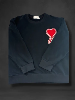 Svart sweatshirt från AMI Paris - Svart sweatshirt från AMI Paris med rund halsringning och ribbade muddar. På bröstet sitter ett stort rött hjärta med bokstaven A, som är typiskt för märket. Tillverkad i mjuk bomullsblandning för skön känsla. Perfekt för en avslappnad och trendig look. Den är M men passar S