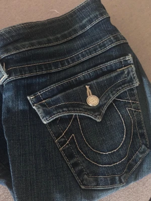 True Religion bootcut jeans blå - Säljer ett par blå bootcut jeans från True Religion med ikoniska kontrastsömmar och snygga detaljer på bakfickan. Jeansen har klassisk femficksdesign och glitter silverfärgade knappar. Perfekta för dig som gillar street och premium denim.