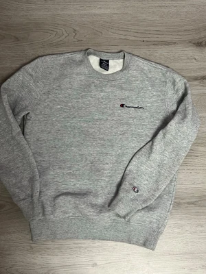 Grå sweatshirt från Champion - Grå melerad sweatshirt från Champion med broderad logga på bröstet och ärmen. Klassisk rund halsringning och ribbade muddar vid ärmslut och nederkant. Mjuk insida för extra komfort, perfekt för en avslappnad stil.